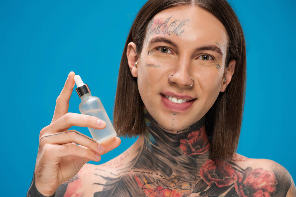 happy and tattooed man holding bottle with serum isolated on blue - Photo, Image