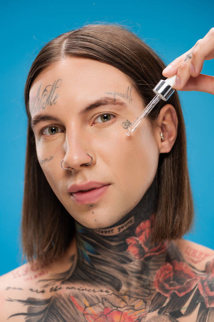jeune homme tatoué appliquant sérum hydratant isolé sur bleu - Photo, image
