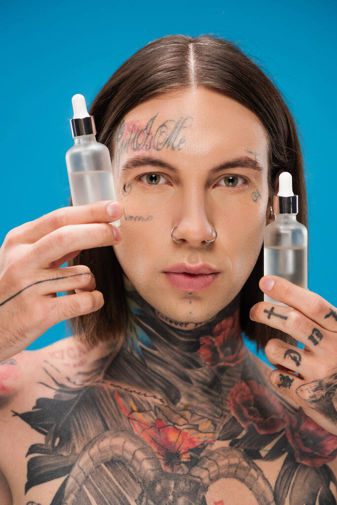 young and tattooed man holding bottles with moisturizing serum isolated on blue - Photo, Image