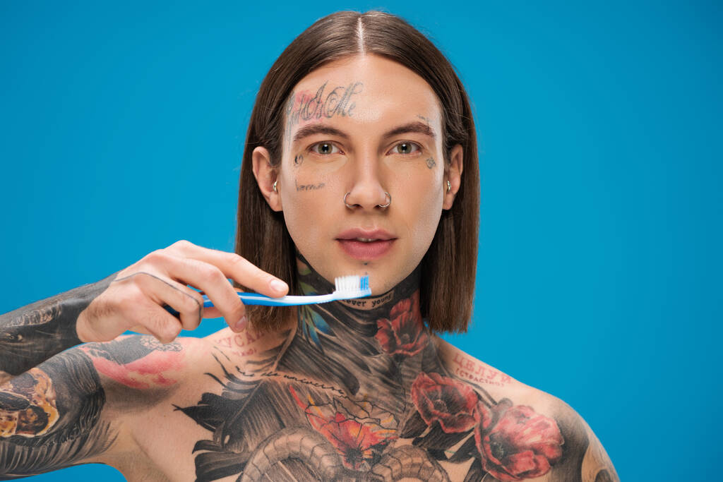 Молодой и татуированный мужчина держит зубную щетку изолированной на голубом - Фото, изображение