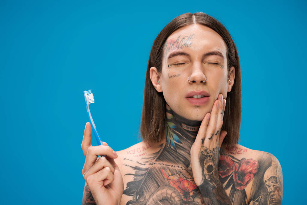 молодий татуйований чоловік із закритими очима тримає зубну щітку ізольовано на синьому
 - Фото, зображення