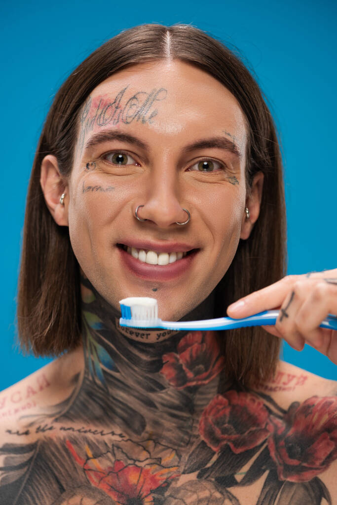 homem positivo e tatuado segurando escova de dentes enquanto sorrindo isolado no azul - Foto, Imagem