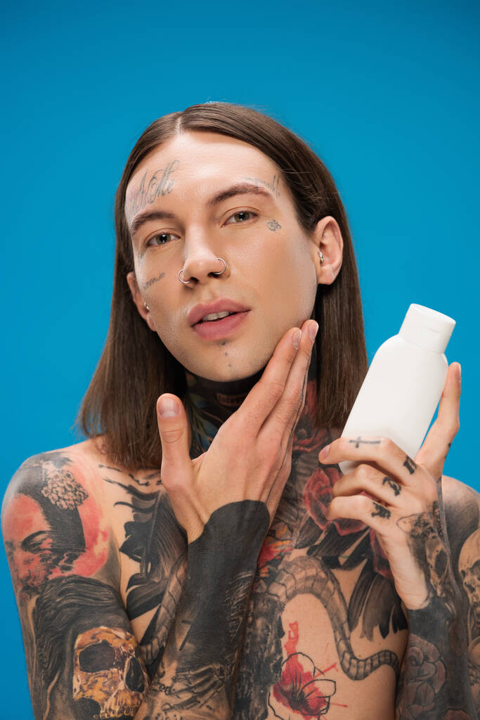 татуированный мужчина держит бутылку с косметическим продуктом и прикосновение гладкой кожи изолированы на синий  - Фото, изображение