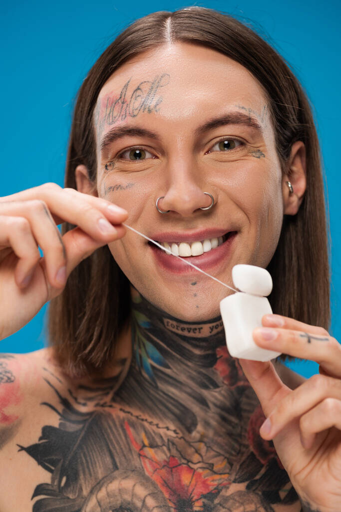 Χαρούμενος και με τατουάζ άντρας που κρατάει οδοντικό νήμα απομονωμένο στο μπλε - Φωτογραφία, εικόνα