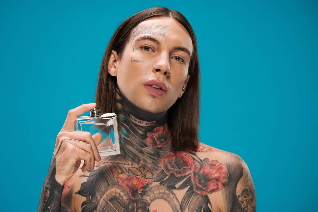nuori ja tatuoitu mies, jolla pullo ja suihkuttava hajuvesi eristetty sinisellä - Valokuva, kuva
