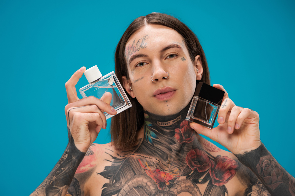 hombre joven y tatuado sosteniendo botellas con perfume aislado en azul - Foto, imagen