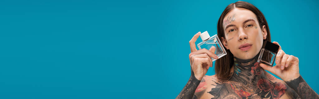 young and tattooed man holding bottles with perfume isolated on blue, banner - Photo, Image