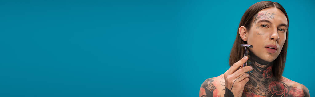 Jongeman met tattoos met scheermes en scheergezicht geïsoleerd op blauw, spandoek - Foto, afbeelding