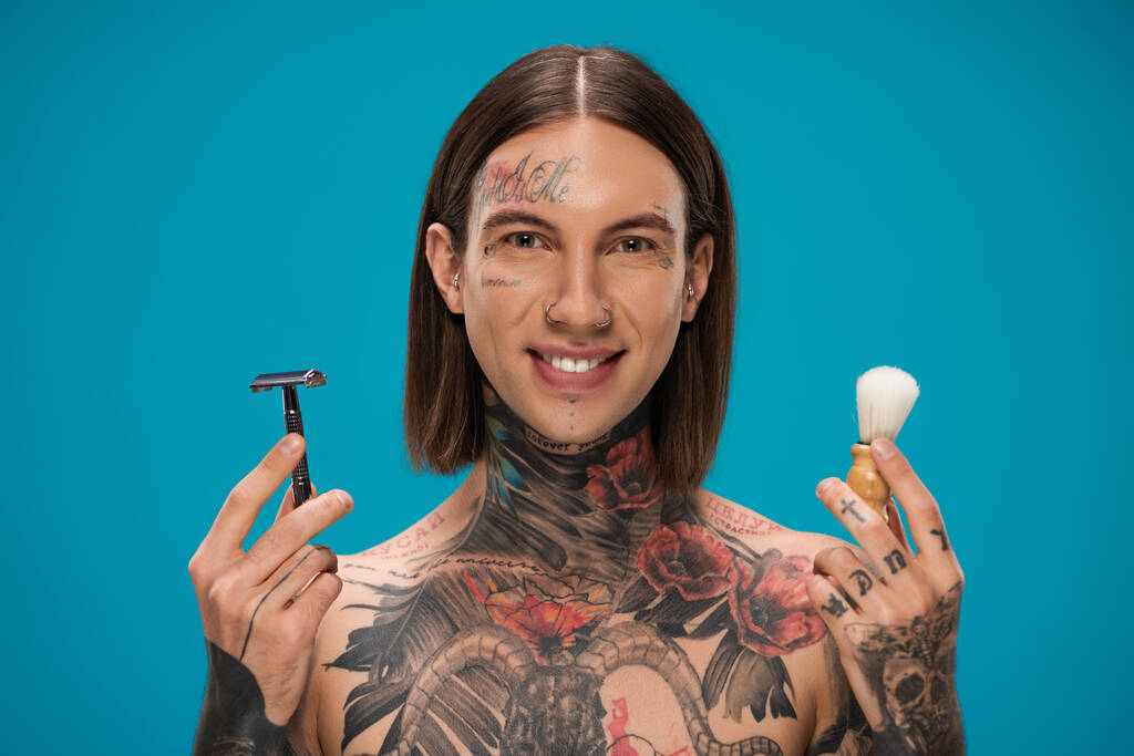 щасливий молодий чоловік з татуюваннями, що тримає запобіжну бритву та вінтажну щітку для гоління ізольовано на синьому
 - Фото, зображення