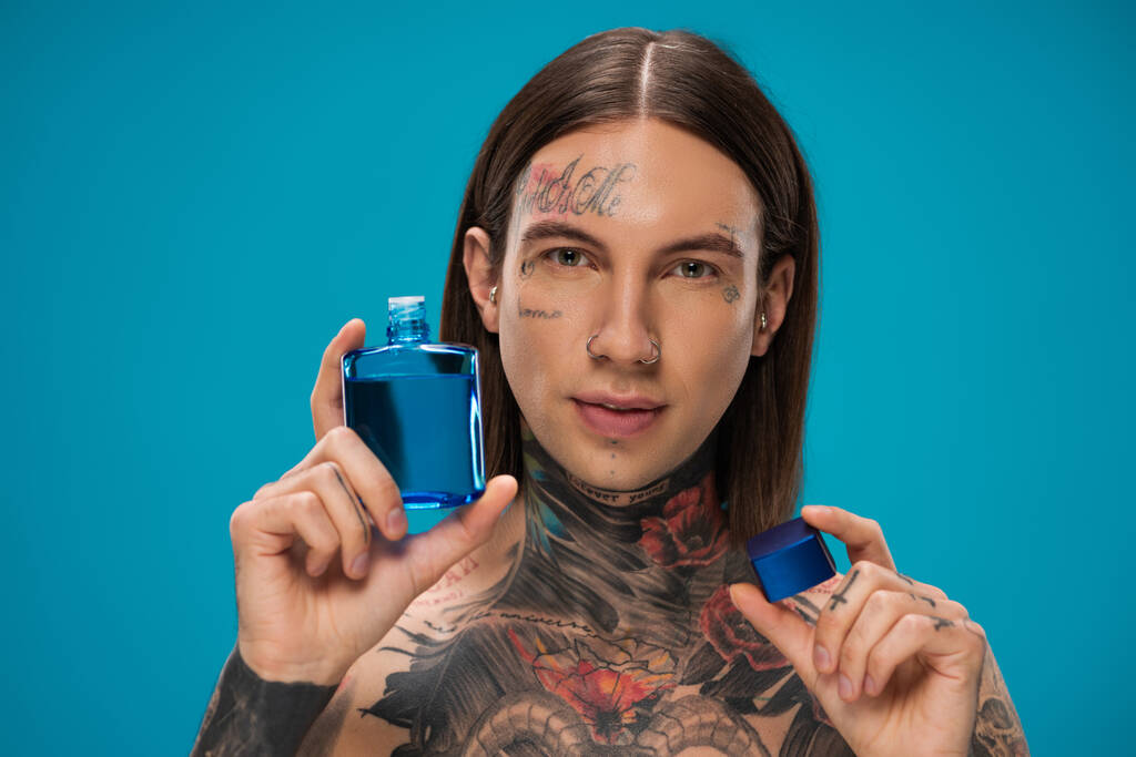τρυπημένος νεαρός άνδρας με τατουάζ κρατώντας μπουκάλι με after shave απομονώνονται σε μπλε  - Φωτογραφία, εικόνα
