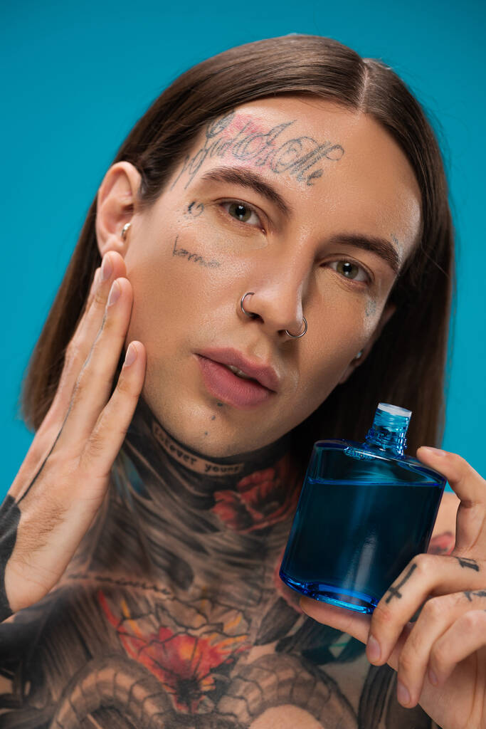 young man with tattoos applying after shave on cheek isolated on blue  - Photo, Image