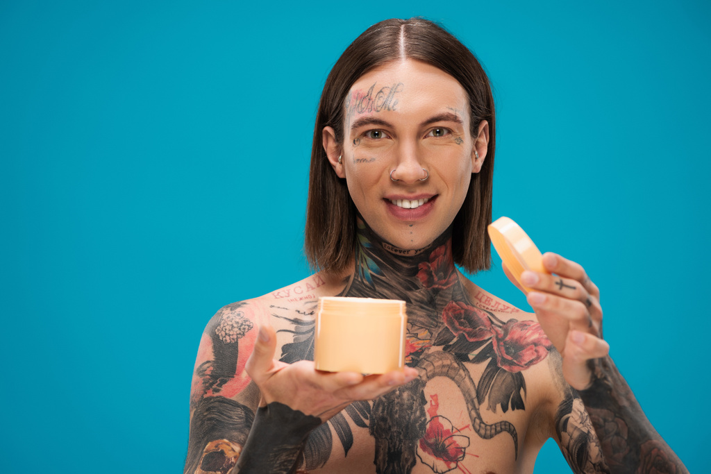 happy young man with tattoos holding container with body butter isolated on blue  - Photo, Image
