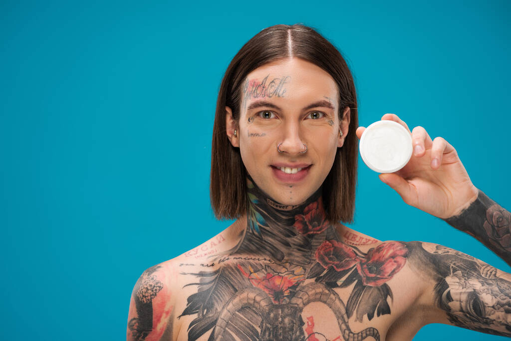 happy young man with tattoos holding container with face cream isolated on blue  - Photo, Image