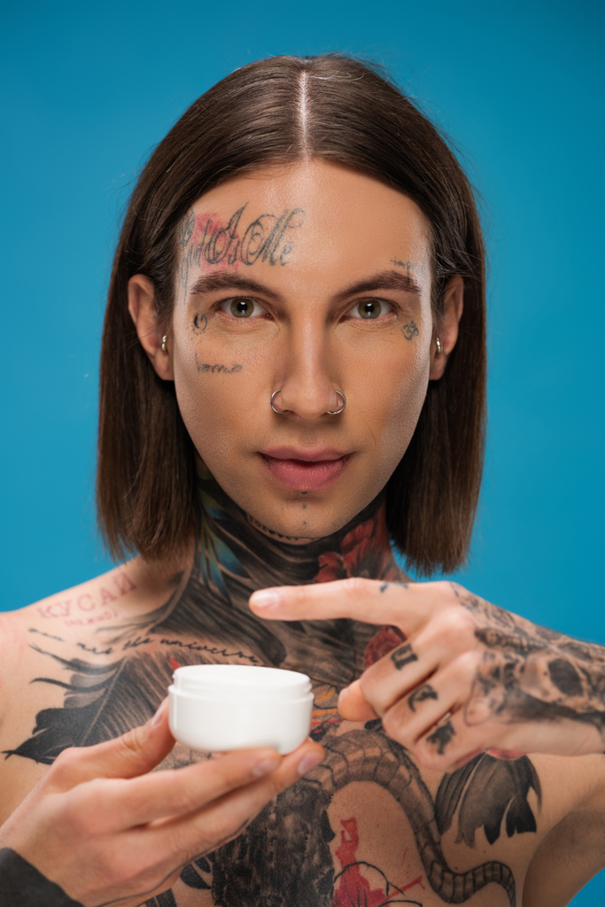 Татуированный молодой человек держит контейнер с кремом для лица изолирован на синий  - Фото, изображение