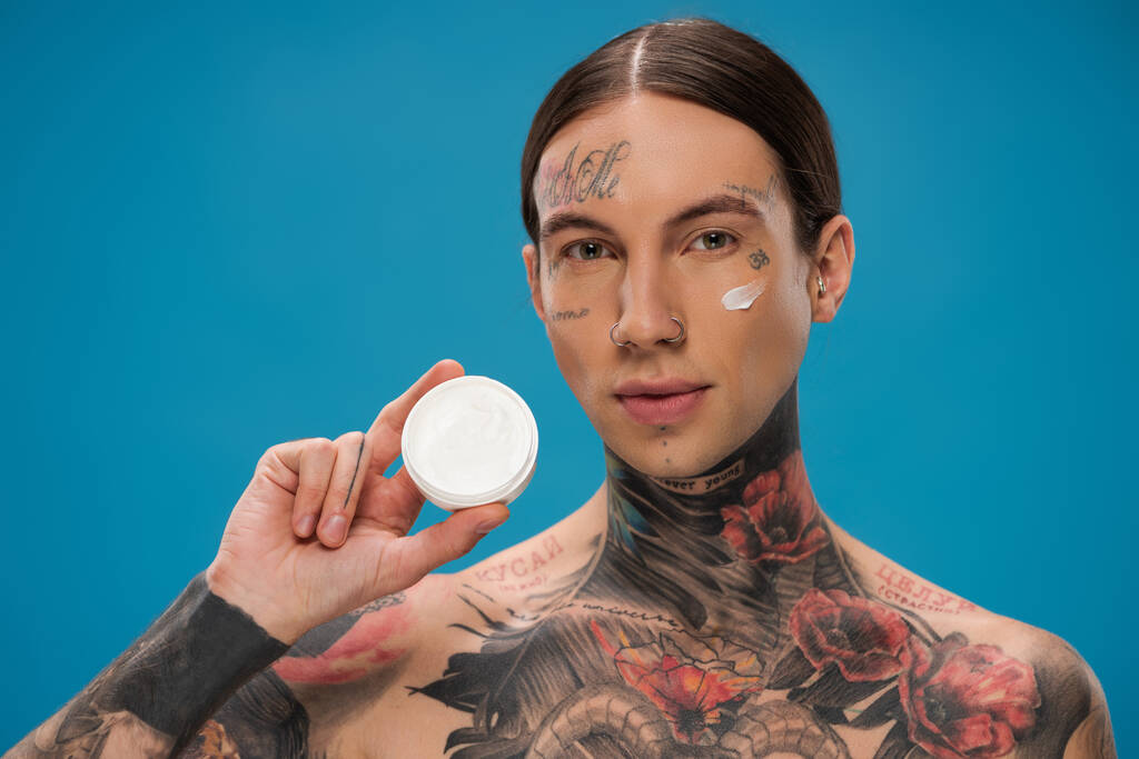 jonge man met tatoeages en crème op wang bedrijf container geïsoleerd op blauw - Foto, afbeelding