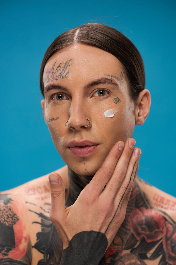 Τατουάζ νεαρός άνδρας εφαρμογή κρέμα στο πρόσωπο και κοιτάζοντας κάμερα απομονωμένη σε μπλε  - Φωτογραφία, εικόνα