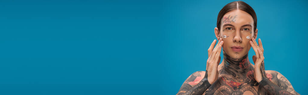 tetovált fiatalember alkalmazása krém az arcon, miközben néz kamera elszigetelt kék, banner - Fotó, kép
