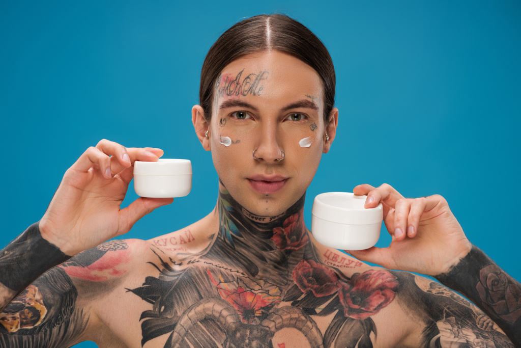 tetovált fiatalember gazdaság különböző tartályok kozmetikai krém elszigetelt kék  - Fotó, kép