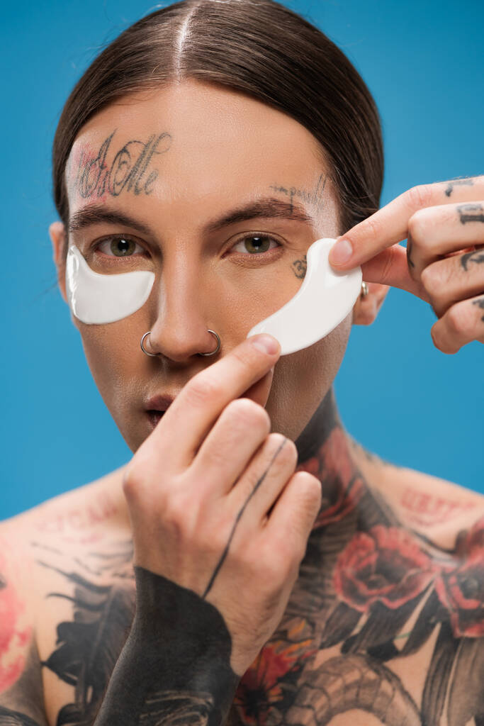 tattooed young man applying eye patches and looking at camera isolated on blue - Photo, Image