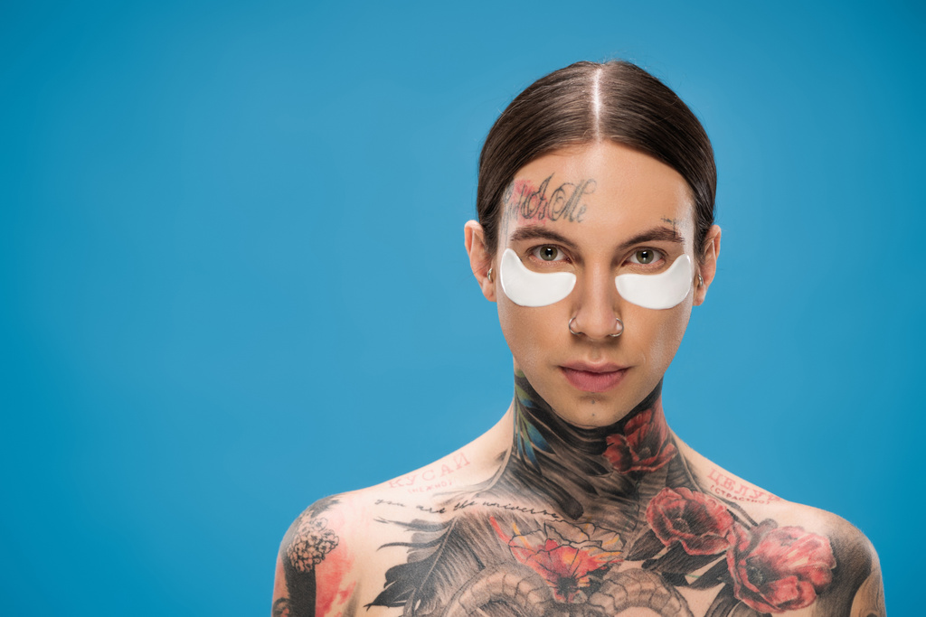 hombre joven sin camisa y tatuado con parches en los ojos mirando a la cámara aislada en azul - Foto, imagen