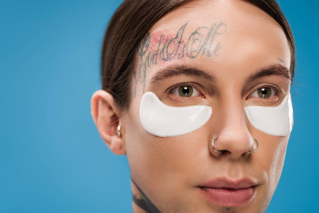 close up of tattooed young man with eye patches looking away isolated on blue - Photo, Image