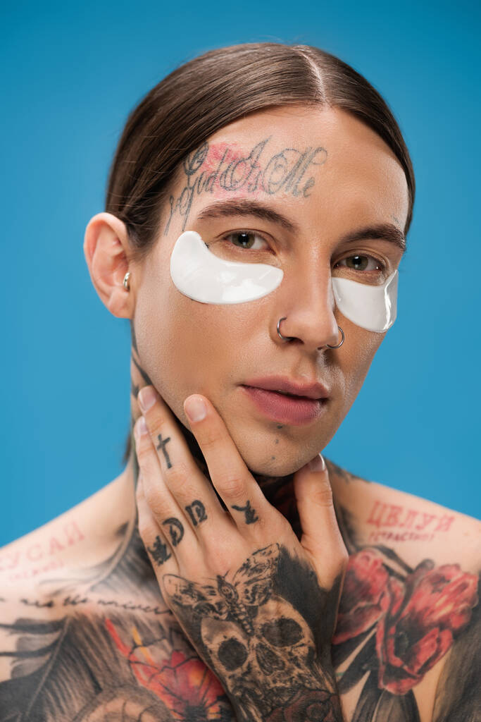 gros plan de jeune homme tatoué avec des patchs pour les yeux regardant la caméra tout en touchant le menton isolé sur bleu - Photo, image