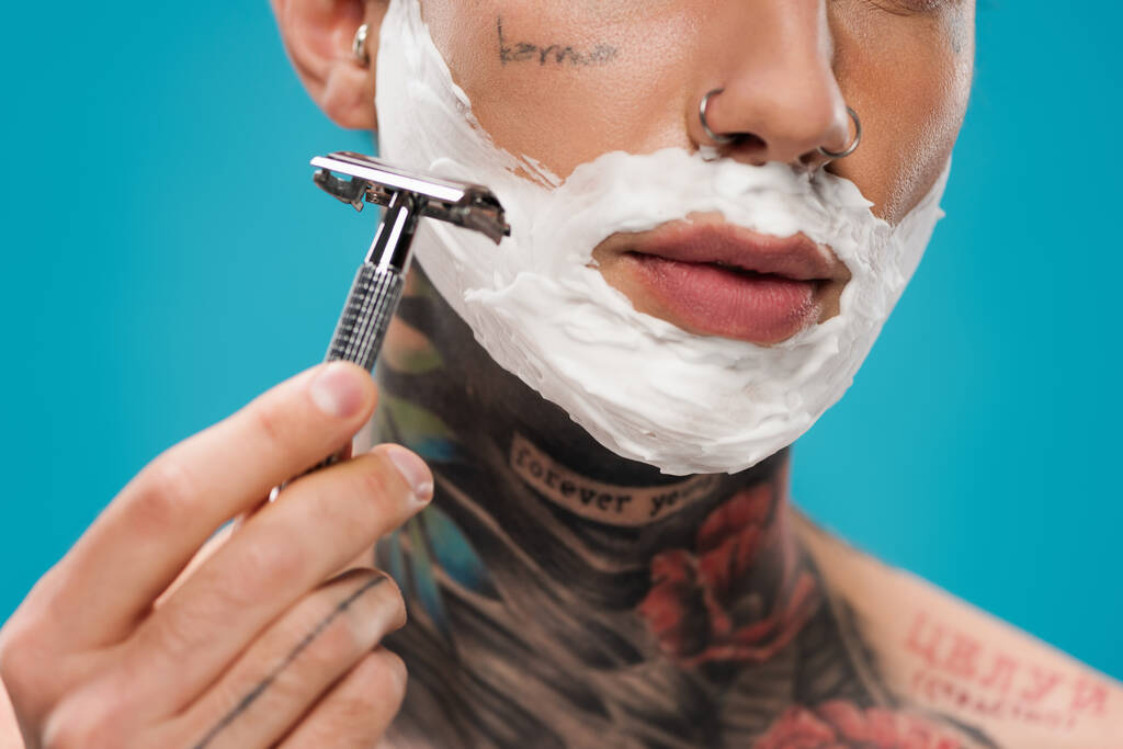 vista recortada de hombre joven tatuado con espuma de afeitar en la cara que sostiene la máquina de afeitar de seguridad aislado en azul  - Foto, imagen