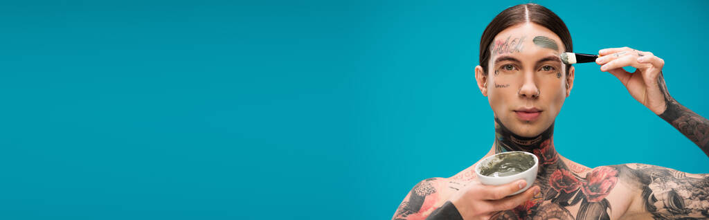 jeune homme tatoué appliquant masque d'argile avec pinceau cosmétique et bol de maintien avec produit isolé sur bleu, bannière - Photo, image