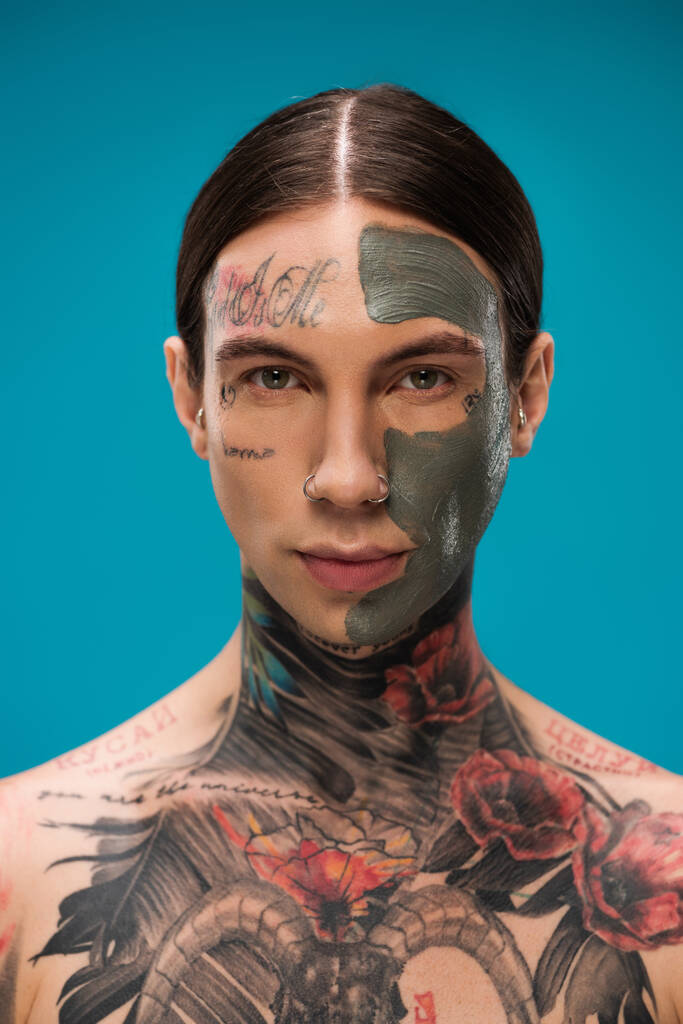 doorboorde jongeman met tatoeages en kleimasker op gezicht kijkend naar camera geïsoleerd op blauw  - Foto, afbeelding