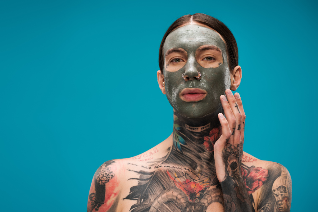 hombre joven sin camisa con tatuajes tocando la cara con máscara de arcilla aislado en azul  - Foto, Imagen