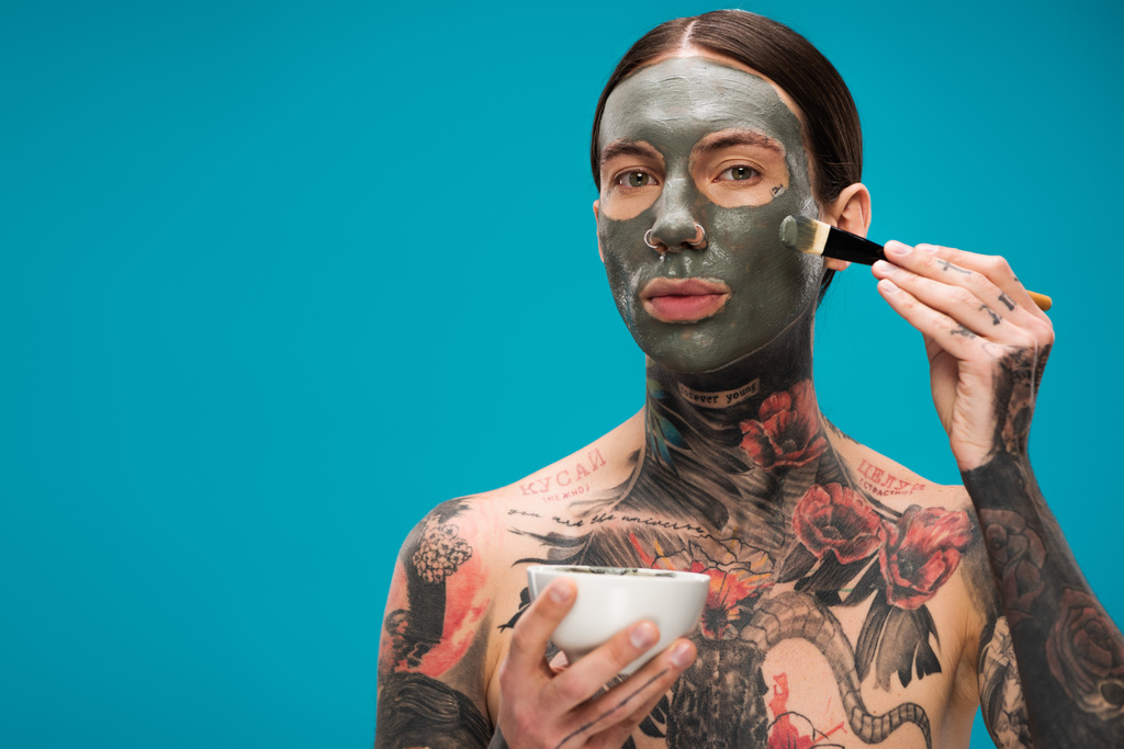Молодой человек без рубашки с татуировками надевает глиняную маску с косметической щеткой и держит миску изолированной на голубом  - Фото, изображение