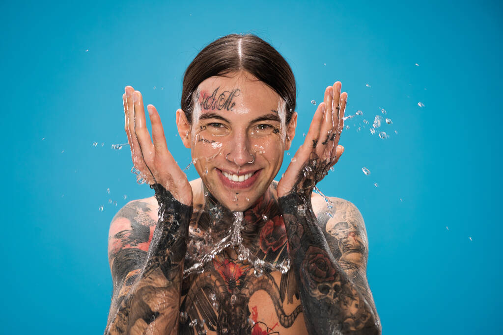Wasserspritzer in der Nähe von fröhlichen Mann mit Tätowierungen Waschen Gesicht isoliert auf blau - Foto, Bild