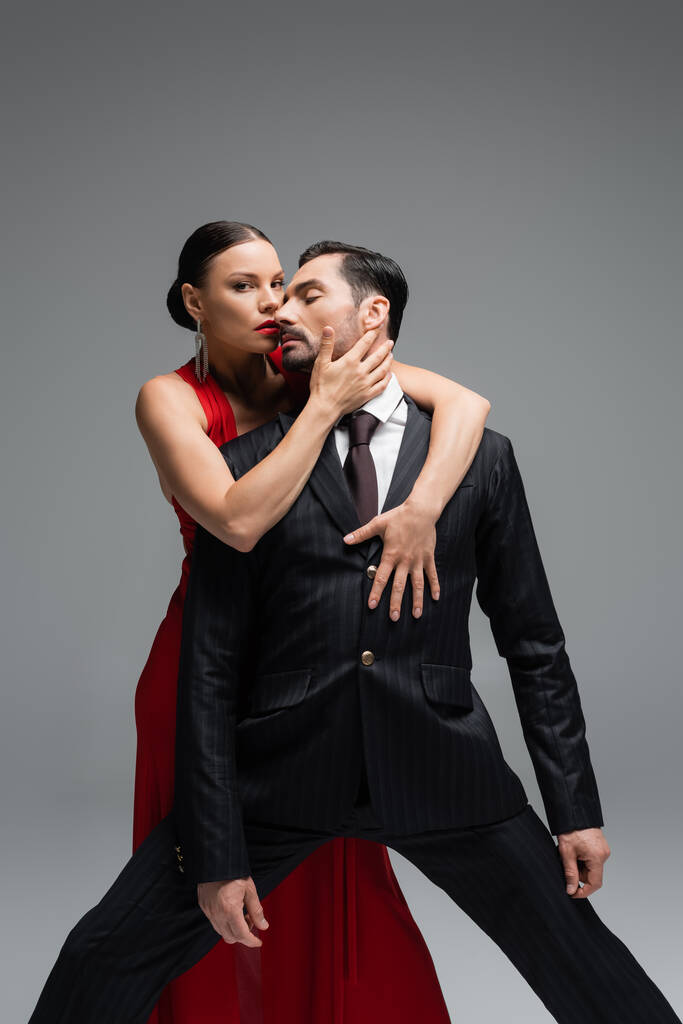 Femme élégante en robe rouge touchant l'homme en costume et regardant la caméra isolée sur gris  - Photo, image
