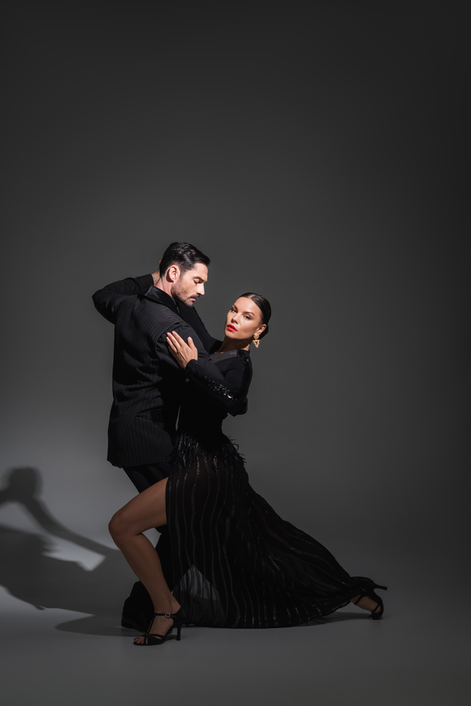 Elegante Frau mit roten Lippen blickt in die Kamera, während sie Tango mit Partner auf grauem Hintergrund mit Schatten tanzt  - Foto, Bild