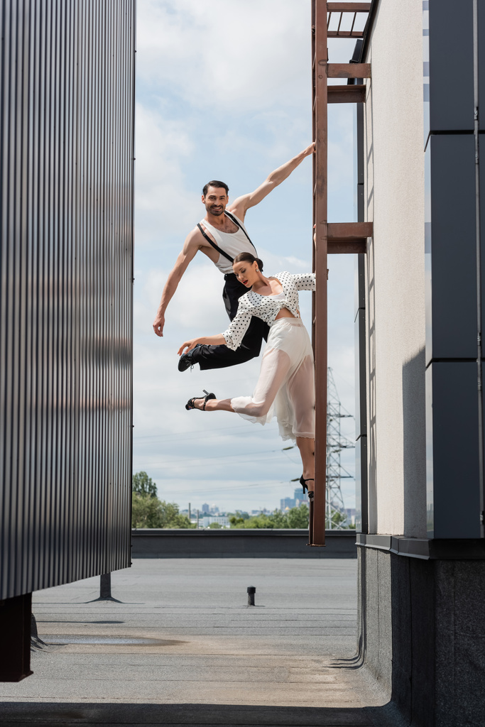 Lächelnde Tänzerin blickt in die Kamera, während sie neben ihrem Partner in Rock und High Heels auf einer Leiter auf dem Dach posiert - Foto, Bild
