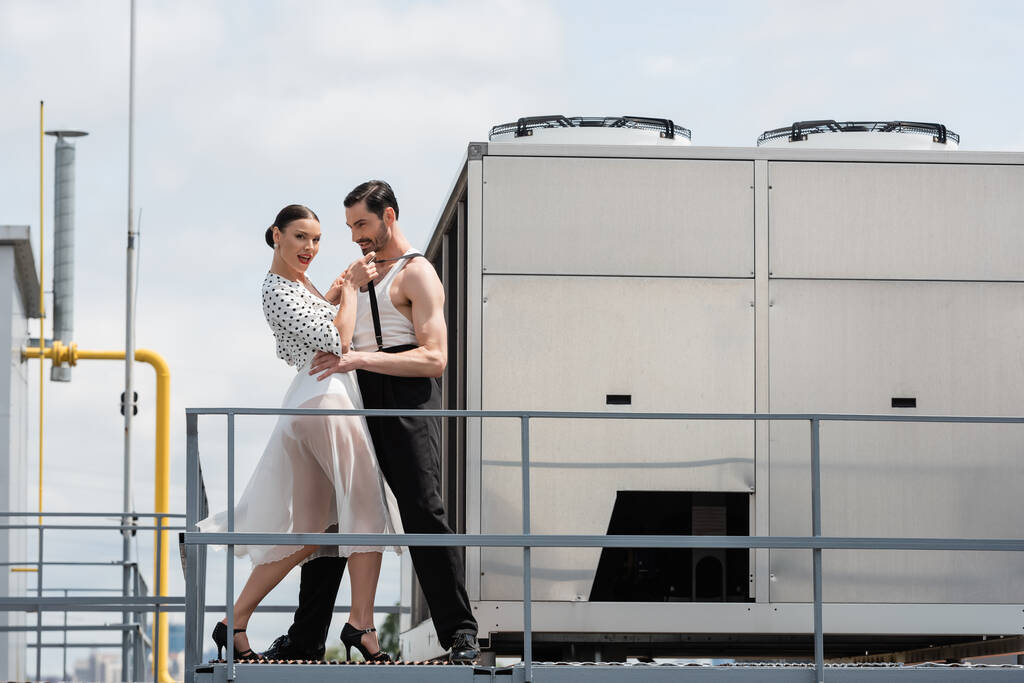 Elegante ballerina professionista in tacchi guardando la fotocamera mentre si muove con il partner sul tetto  - Foto, immagini