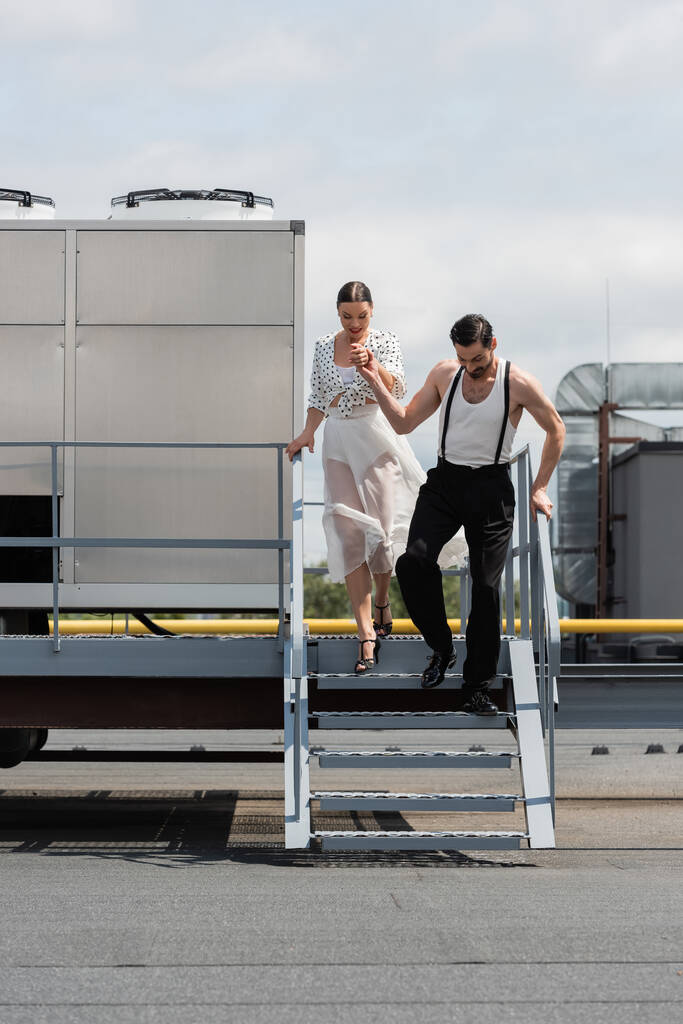 Elegante par de bailarines tomados de la mano mientras caminan por las escaleras en el techo  - Foto, imagen