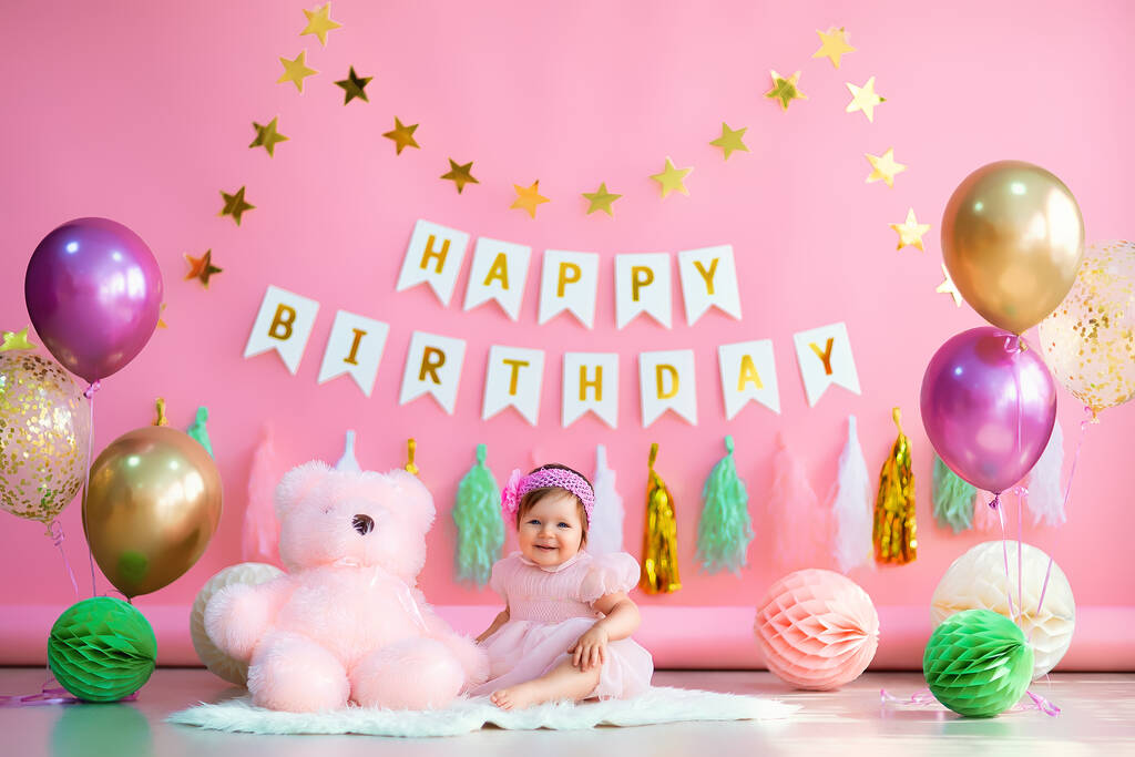 かわいい赤ちゃんの女の子は、手紙でピンクの背景にピンクの休日のドレスでピンクのテディと紙のボールとカラフルな空気の風船の近くに座っています。誕生日おめでとう - 写真・画像