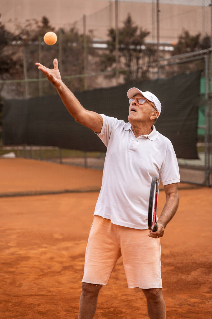 Senior tennisspeler gekleed in sportkleding in actie op een tennisbaan van klei - gepensioneerd wellness concept - Foto, afbeelding