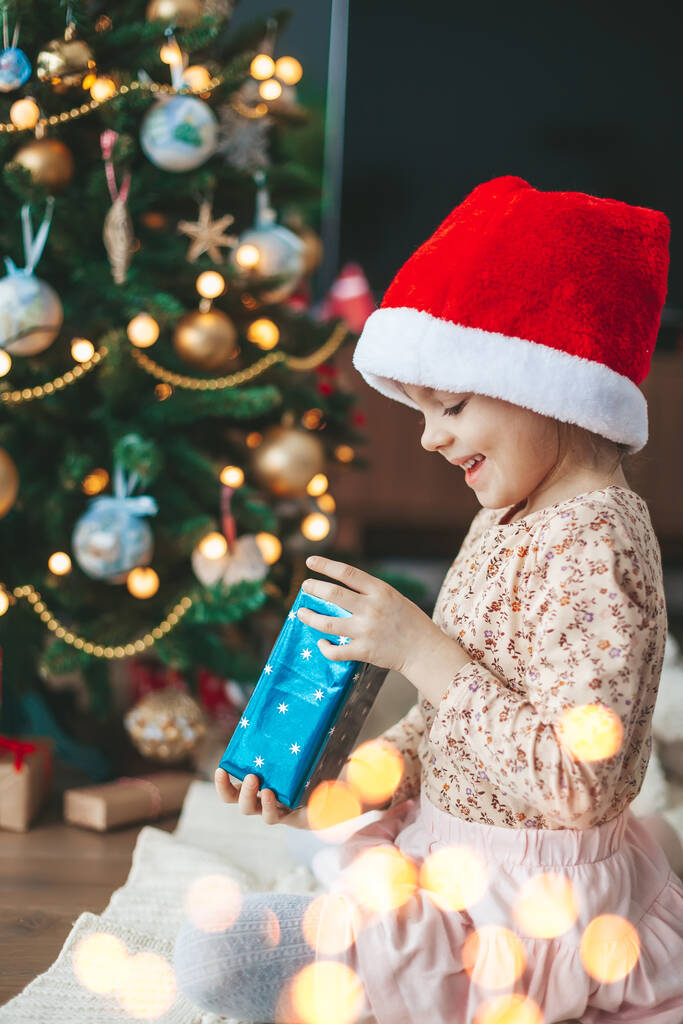 Sorrindo menina bonito em chapéu vermelho Papai Noel abrir uma caixa de presente sentado perto da árvore de Natal. Feliz Natal e Feliz Ano Novo conceito. - Foto, Imagem