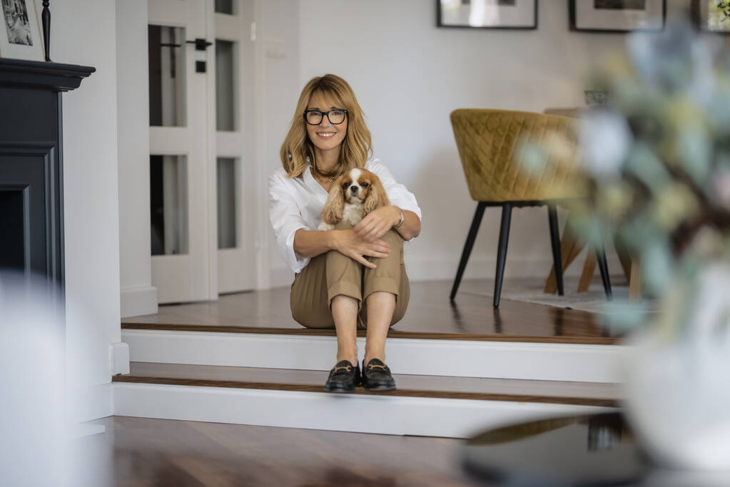 Full length of happy middle aged woman sitting on the floor white her cute puppy. Attractive female wearing glasses and casual clothes while relaxing at home. - Photo, Image