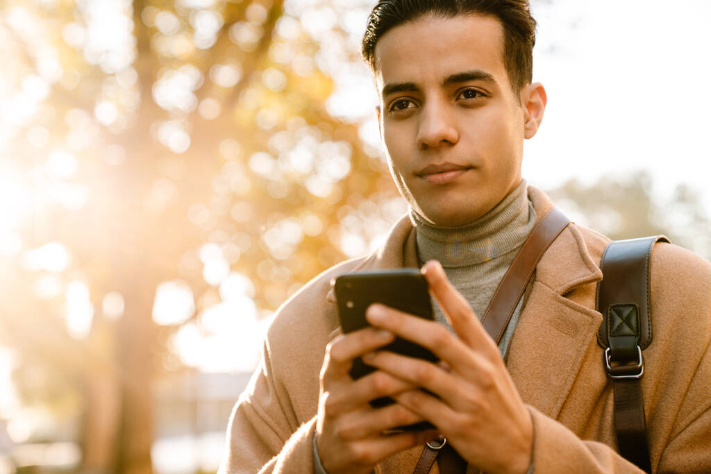 Young man wearing coat using mobile phone while walking in park outdoors - Photo, Image