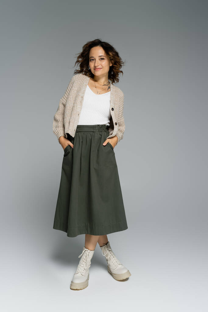 full length of young woman in cozy cardigan and boots posing with hands in pockets of green skirt on grey background - Photo, Image