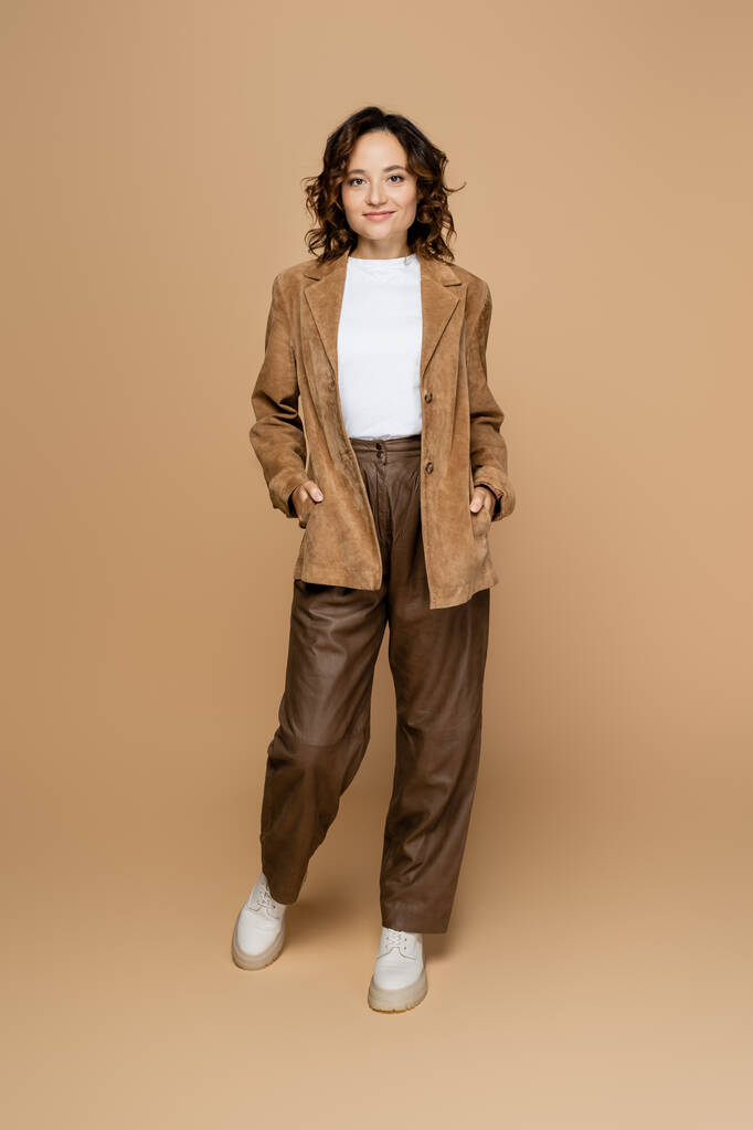 longitud completa de la mujer morena de pie con las manos en bolsillos de gamuza chaqueta marrón sobre fondo beige - Foto, Imagen