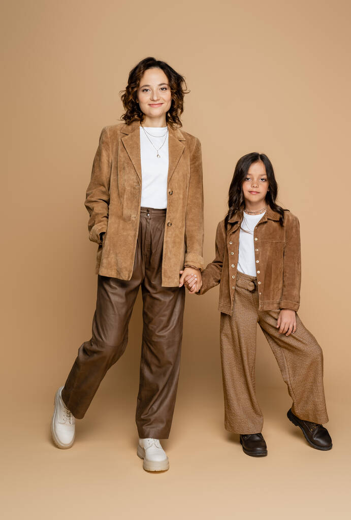 longitud completa de la mamá y la hija en pantalones marrones y chaquetas de gamuza cogidas de la mano sobre fondo beige - Foto, Imagen