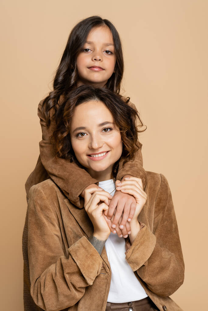 smiling woman in brown suede jacket holding hands of daughter and looking at camera isolated on beige - Photo, Image