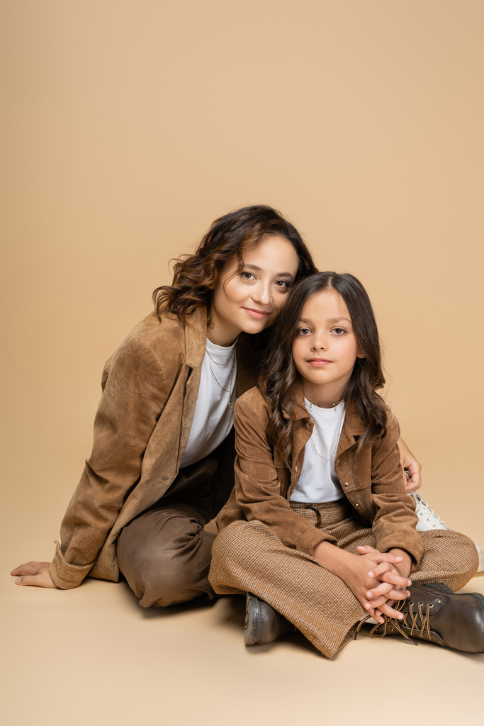 moeder en dochter in suède jassen en bruine broek glimlachen op camera terwijl zitten op beige achtergrond - Foto, afbeelding