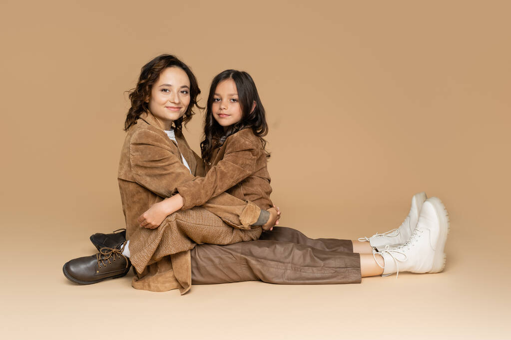šťastná žena a dívka objímající zatímco sedí v semišové bundy a boty na béžovém pozadí - Fotografie, Obrázek