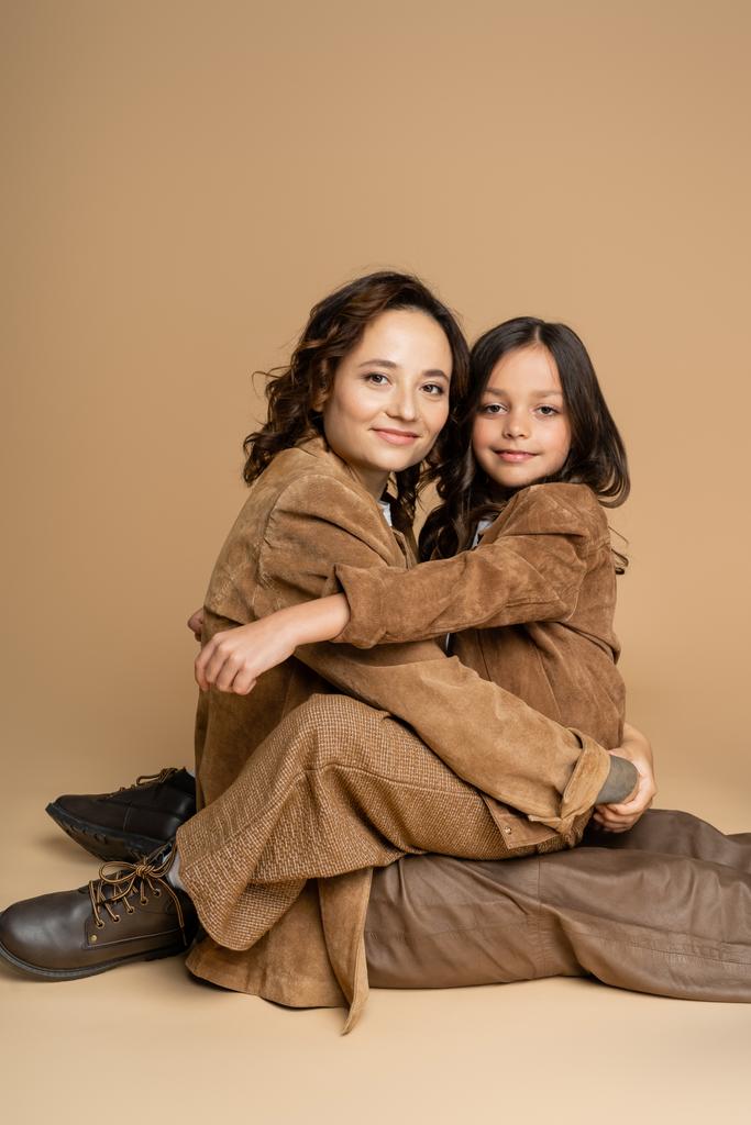 moeder en dochter in suède jassen en bruine broek glimlachen op camera terwijl zitten op beige achtergrond - Foto, afbeelding