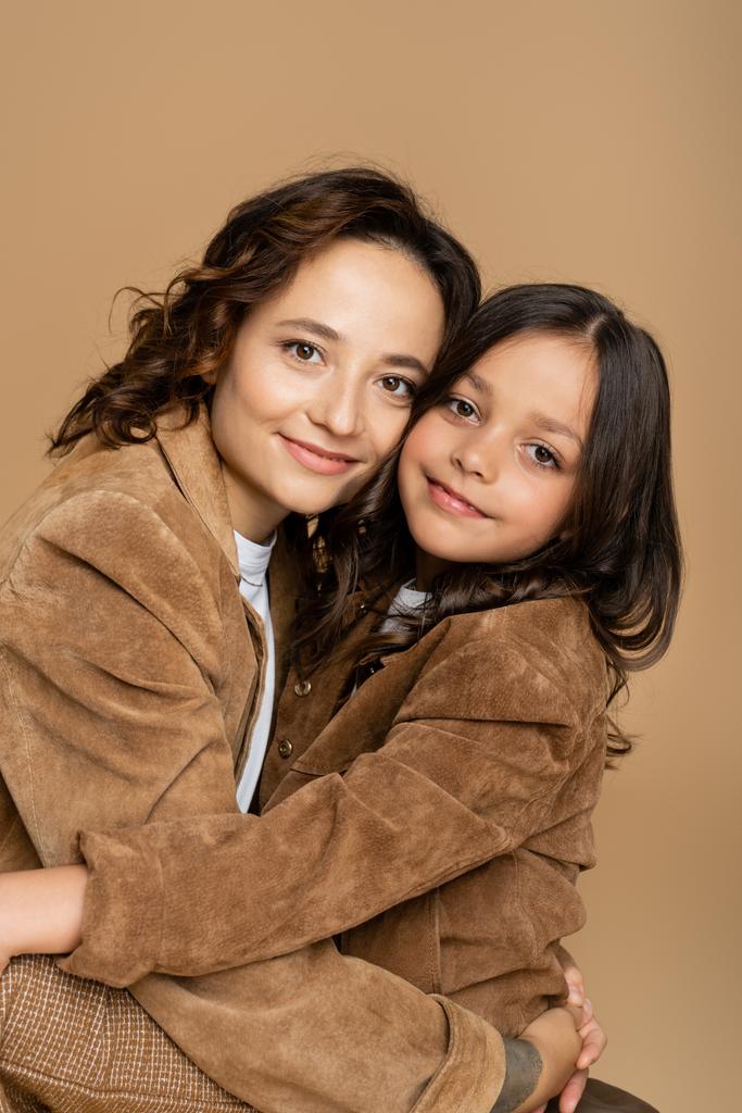 ブルネット母と娘で茶色のジャケット抱擁と笑顔でカメラ絶縁上ベージュ - 写真・画像